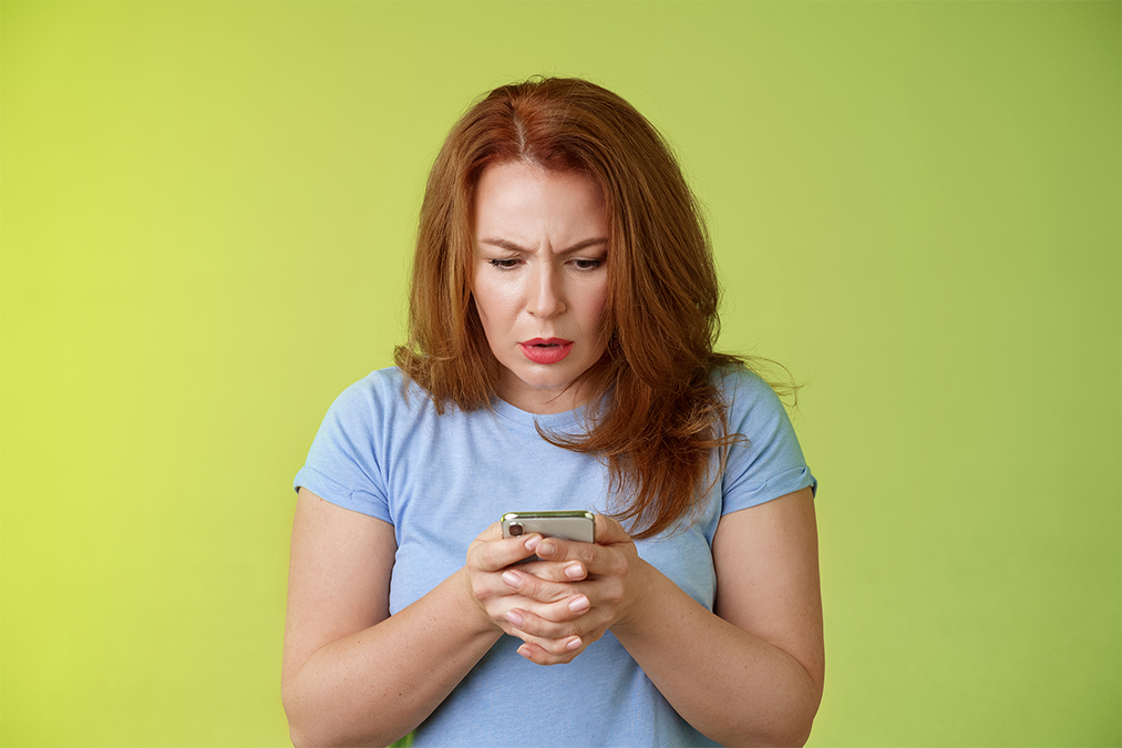 Portrait Lifestyle. Confused unsure redhead middle-aged woman learn how use social media trying understand emoji look intense focused smartphone display reading important news message stand green background.